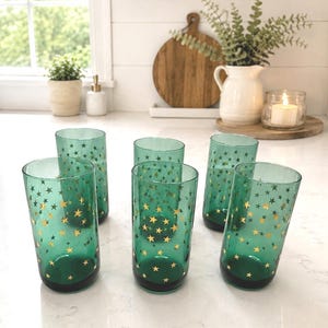 May include: A set of five cylindrical drinking glasses made of translucent green glass, each decorated with a repeating pattern of small, metallic gold stars scattered across the exterior surface of the glass walls.