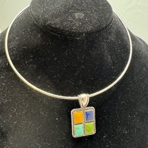 May include: A silver-toned choker necklace with a square pendant. The pendant features four colorful squares: orange, blue, green, and turquoise. The pendant is framed with a twisted rope design.