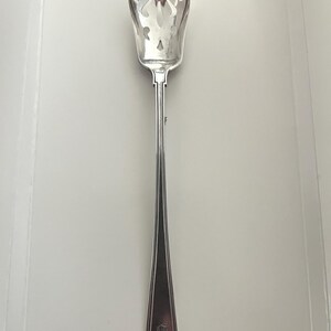 May include: A silver-toned metal serving spoon featuring a pointed, perforated bowl with a decorative cutout pattern. The long, slender handle has a tapered design with a flat, squared-off end and an engraved monogram detail near the base.