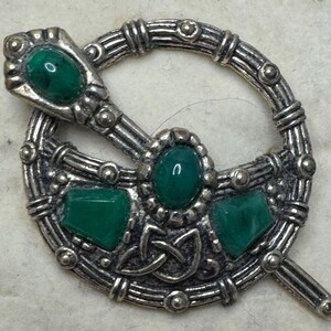 May include: A silver-toned brooch with a Celtic knot design. The brooch features oval and square green gemstones. The pin is a circular shape with a pin that goes through the circle. The brooch has a vintage look.