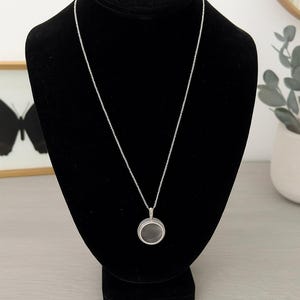 May include: A silver necklace with a circular pendant is displayed on a black mannequin. The pendant has a dark gray center and a silver frame. The necklace chain is delicate and hangs down the mannequin's chest.