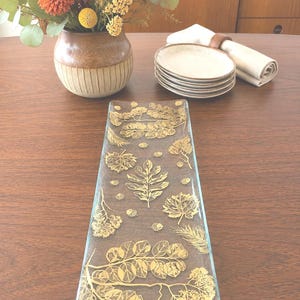 May include: A rectangular glass serving tray featuring a textured surface with a gold-colored botanical pattern of various leaves, branches, and small acorns scattered across the clear glass. The tray has a smooth, flat design with straight edges.