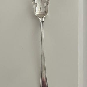 May include: A silver-toned metal serving spoon featuring a perforated, shield-shaped bowl with decorative cutouts. The long, slender handle has a smooth, tapered design that widens at the base, which is finished with a geometric Greek key pattern.