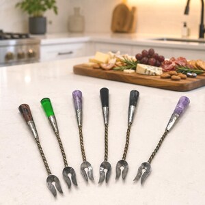 May include: A set of six appetizer forks with twisted metal stems and decorative handles in various colors including brown, green, purple, and black. Each fork features a two-pronged head and a silver-toned metal ferrule connecting the handle.