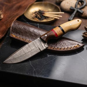 May include: A Damascus steel knife with a wooden handle featuring red and black accents, resting in a brown leather sheath. The blade has a unique pattern. The knife is displayed on a black marble surface.
