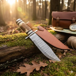 Könnte beinhalten: Ein Jagdmesser mit gemusterter Klinge und einem Griff mit hellen und dunklen Brauntönen, das auf einem moosbedeckten Baumstamm liegt. Eine braune Lederscheide befindet sich neben dem Messer. Der Hintergrund umfasst eine Ledertasche, einen Kompass und eine Karte.
