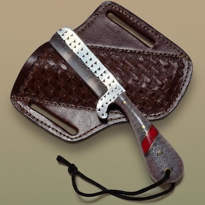 May include: A straight razor with a silver blade featuring triangular cutouts, resting on a brown leather sheath. The handle has a gray, textured pattern with a red stripe and gold accents. A black cord is attached.