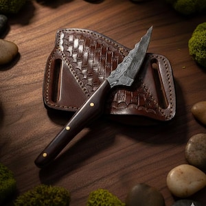 May include: A small, ornate knife with a dark wooden handle and a patterned steel blade rests on a brown leather sheath. The sheath has a woven design. The knife is surrounded by small stones and moss on a wooden surface.