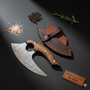 May include: A handcrafted cleaver knife with a patterned blade and a wooden handle, next to a brown leather sheath. The knife has a circular opening near the handle. The words "HANDCRAFTED IN USA" are on a small leather tag.