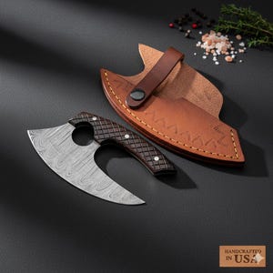 May include: A Damascus steel cleaver with a dark brown, diamond-patterned handle and a leather sheath. The cleaver has a curved blade and a circular cutout. The sheath is brown with a strap and button closure. "Handcrafted in USA" is printed on the bottom right.