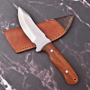 May include: A stainless steel hunting knife with a wooden handle and a brown leather sheath. The knife has a sharp, pointed blade and a curved handle. The sheath is stitched with yellow thread.
