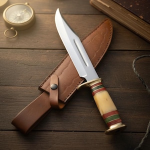 May include: A hunting knife with a polished steel blade and a bone handle accented with red and green stripes. The knife rests in a brown leather sheath on a wooden surface, with a compass and a book in the background.