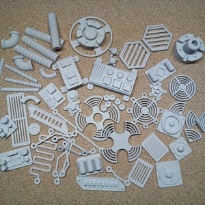 Op de afbeelding: Een verzameling lichtgrijze, ongeverfde 3D-geprinte kunststof onderdelen met diverse mechanische en industriële vormen, waaronder buizen, ventilatieopeningen, ronde roosters, bedieningspanelen en geometrische verbindingsstukken voor modelbouw.