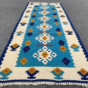 Puede incluir: Una alfombra rectangular tejida con un campo central turquesa con patrones geométricos blancos, amarillos y azules. La alfombra tiene un borde blanco con motivos florales azules y amarillos y un borde con flecos. La alfombra mide aproximadamente 152 cm de largo.