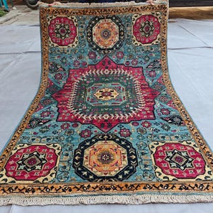 Op de afbeelding: Een rechthoekig vloerkleed met een levendige turquoise achtergrond, een groot centraal magenta medaillon en kleinere achthoekige motieven in magenta, zwart en goud. Het kleed heeft een gedetailleerde rand en witte franjes aan de korte zijden.