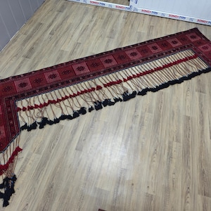 May include: A long, L-shaped textile valance featuring a deep red and black geometric pattern. The bottom edge is adorned with a series of hanging decorative cords, each finished with small red and black tassels, creating a layered fringe effect.