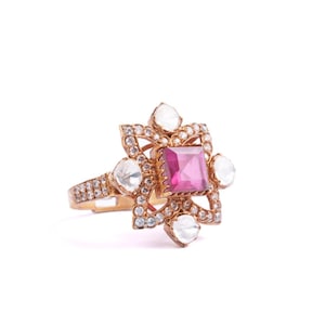 May include: A gold-toned ring featuring a square, pink gemstone at its center, surrounded by a floral design. The ring is embellished with numerous small, clear gemstones and larger, pear-shaped stones.