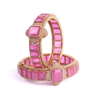 Puede incluir: Dos brazaletes dorados con gemas rosas rectangulares y pequeños acentos blancos. Los brazaletes tienen un diseño de bisagra. Las gemas son de color rosa vibrante, sobre un fondo blanco.