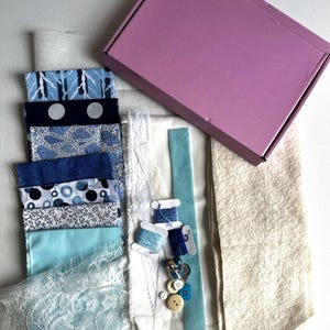 May include: A craft kit containing a rectangular pink cardboard box, a stack of blue and white patterned fabric swatches, a piece of white lace, a strip of light blue ribbon, three spools of blue thread, small buttons, and a piece of beige felt.