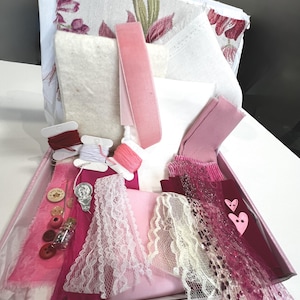 May include: A craft kit containing pink and white fabrics, lace, ribbon, and thread spools. The kit includes buttons, a small glass bottle, and a decorative pin. The fabrics feature floral patterns and various textures.