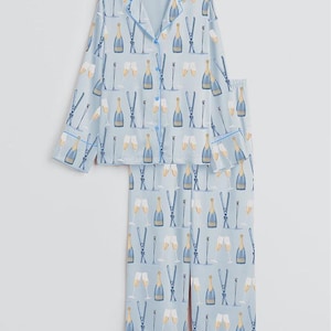 May include: Light blue pajama set with a repeating pattern of champagne bottles, glasses, and skis. The long-sleeved top has a button-down front and blue trim. The pants are straight-legged with matching trim. A fun and festive sleepwear set.