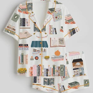 May include: White pyjama set with a book-themed design. The short-sleeved top and shorts feature illustrations of books, open books, and a bookstore. The set has yellow trim and a button-down front.
