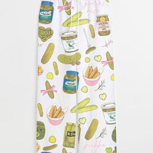 May include: White pajama pants with a repeating pattern of pickles, pickle jars, and pink bows. The text "pickbe girlies rise" is printed in pink script on the pants. The pants have a drawstring waist.