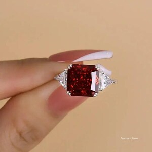 May include: A silver-toned ring featuring a large, square-cut, deep red gemstone at the center, flanked by two smaller, triangular-cut clear white stones on each side. The setting is metallic, and the text Ammar Gems is visible in the corner.