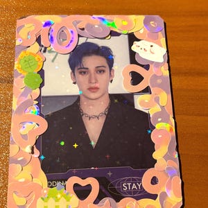 Puede incluir: Una tarjeta fotográfica rectangular con una persona con cabello azul y una parte superior negra, enmarcada por un borde rosa iridiscente con formas decorativas y la palabra "STAY". La tarjeta tiene un efecto holográfico con acentos en forma de estrella.