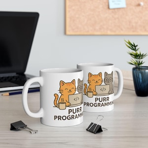Pode incluir: Duas canecas de cerâmica branca com um desenho de desenho animado de gatos laranja e cinza programando em laptops. As canecas têm o texto "PURR PROGRAMMING" impresso. Um laptop preto e material de escritório estão no fundo.