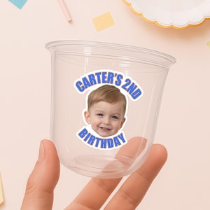 May include: Clear plastic cup with a blue and white sticker that reads "Carter's 2nd Birthday" with a photo of a smiling toddler. The cup is held by a hand against a light pink background with colorful confetti.
