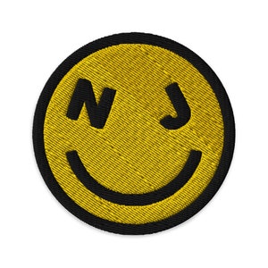 Könnte beinhalten: Ein gelber Smiley-Patch mit schwarzem Rand und den Buchstaben "NJ" für New Jersey in Schwarz.