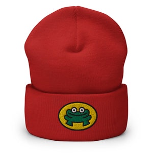 Puede incluir: Gorro de punto rojo con una rana verde bordada en la parte delantera. La rana sonríe y tiene ojos amarillos.