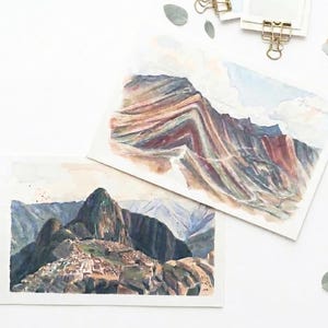 May include: Two watercolor paintings on white paper. One depicts the colorful, striped layers of Vinicunca mountain in shades of red, gold, and teal. The other shows the stone ruins of Machu Picchu nestled among dark, craggy mountain peaks.