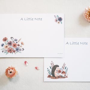 May include: Two white rectangular note cards with the text A Little Note. One card features a watercolour floral bouquet in shades of orange, blue, and green. The other card displays a watercolour illustration of a hedgehog with a mushroom.