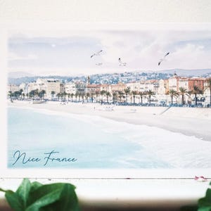 May include: A rectangular art print featuring a coastal scene of Nice, France, with a sandy beach, turquoise water, palm trees, and buildings in the background. Three birds fly in the pale sky. The text Nice France is written in blue script.