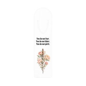 Könnte beinhalten: Weißer Lesezeichen mit einem Schwert, das mit rosa und pfirsichfarbenen Rosen verziert ist. Der Lesezeichen hat den Text "You do not fear. You do not falter. You do not yield." in schwarzer, gotischer Schrift. Ein dekorativer Artikel für Bücherliebhaber.