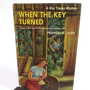 May include: A vintage paperback book cover with a yellow title "WHEN THE KEY TURNED" and the subtitle "Danger follows Kay as she searches for a priceless violin". The author is Frances K. Judd. The cover art features a woman in a blue and red dress sitting on a porch with a wooden chest in front of her. Two men are standing in the doorway behind her.