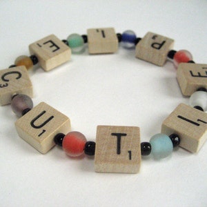 Op de afbeelding: Een armband gemaakt van houten lettertegels die het woord "CUTE" spellen met zwarte en witte kralen ertussen. De tegels zijn bruin en de kralen zijn zwart en wit.