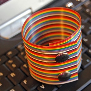 May include: A colorful rainbow-striped bracelet with two silver snaps. The bracelet is made from a flat, flexible material and is lying on a black laptop keyboard.