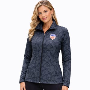 May include: A dark blue zip-up jacket with a subtle geometric pattern. The jacket has a stand-up collar and a small US emblem on the chest. The jacket is worn with black trousers.