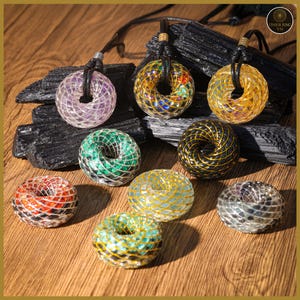 May include: A collection of handcrafted crystal pendants in various colors, including purple, green, gold, and red. Each pendant features a unique design with a woven metal cage and a black cord. The pendants are displayed on a wooden surface with black stone accents.
