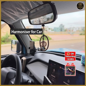 May include: Interior car shot featuring a rearview mirror and a decorative brown pendant hanging from it. The pendant has an oval shape with a spherical design inside. Text on the image reads "Harmoniser for Car". A gift box with the text "Tensor Ring 144 MHz" is also visible.