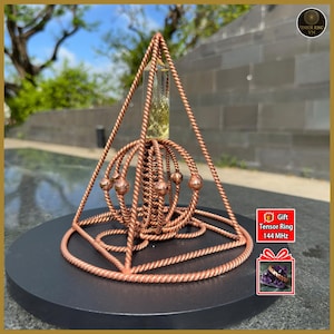 May include: A copper-coloured geometric sculpture featuring a pyramid shape. The sculpture has a spherical design with small copper spheres. It sits on a black circular platform. The image includes the text "Tensor Ring VN" and "Gift Tensor Ring 144 MHz".