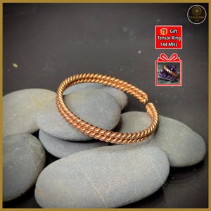 May include: A copper-colored bracelet with a twisted rope design, resting on gray stones. The bracelet is a gift, as indicated by the text "Gift Tensor Ring 144 MHz" in a small graphic. The bracelet is a piece of jewelry.