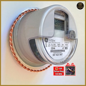 Puede incluir: Un medidor de electricidad con un anillo de color cobre enrollado alrededor. El medidor muestra una lectura digital y tiene varias etiquetas. Una pequeña caja de regalo con texto está en la esquina inferior derecha.