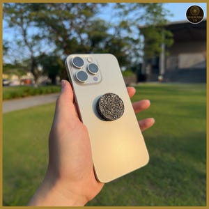 Puede incluir: Un teléfono inteligente dorado con un agarre circular negro para teléfono adjunto a la parte posterior. El agarre del teléfono tiene un diseño geométrico detallado. El teléfono se sostiene en una mano contra un fondo borroso al aire libre.