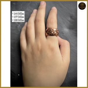 May include: A copper-colored ring with a textured, organic design is worn on a hand. The ring has a wide band and a surface that appears to be made up of overlapping leaf-like shapes. The text "144 Mhz, 177 Mhz, 188 Mhz" is visible.