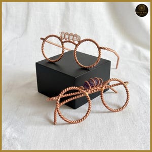 May include: Two pairs of copper-coloured wire-framed glasses with round lenses. One pair features clear crystal accents, the other purple amethyst. The frames are displayed on a black box against a white background.