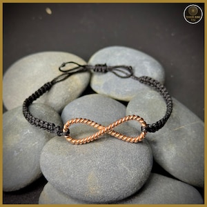 Op de afbeelding: Een zwart macramé armband met een koperkleurig oneindigheidssymbool. De armband wordt getoond op grijze stenen. Het oneindigheidssymbool is gemaakt van gedraaide koperdraad. De armband is verstelbaar met een schuifknoopsluiting.